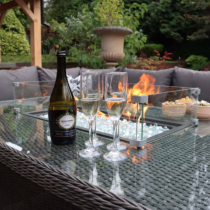 Close-up of the Paris Fire Pit Corner Dining & Lounge Set's glass-top table, featuring a bottle of champagne and glasses next to the warm fire pit, set against lush garden scenery.