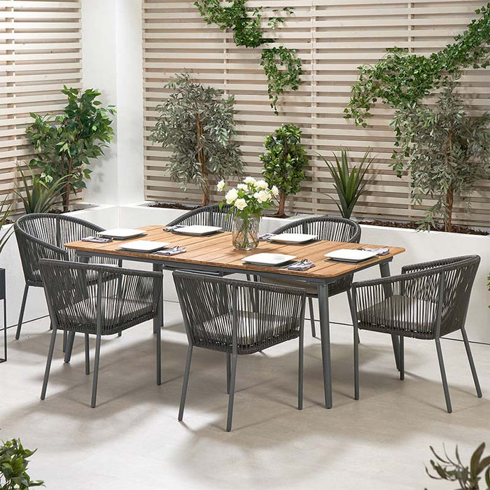 A beautiful Reims Outdoor Dining Set placed in a modern garden setting, featuring a wooden dining table and six chairs with grey polystring weave, surrounded by greenery and decorative elements.