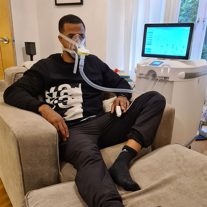ReOxy Hypoxic Therapy Device in use, man in black sweatshirt seated in armchair wearing full face breathing mask and finger pulse oximeter, device screen visible in background, home setting.