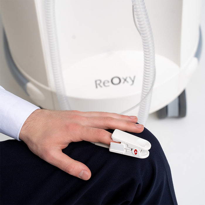 ReOxy Hypoxic Therapy Device pulse oximeter finger clip worn on a hand resting on knee, white ReOxy unit visible in background, clinical setting.