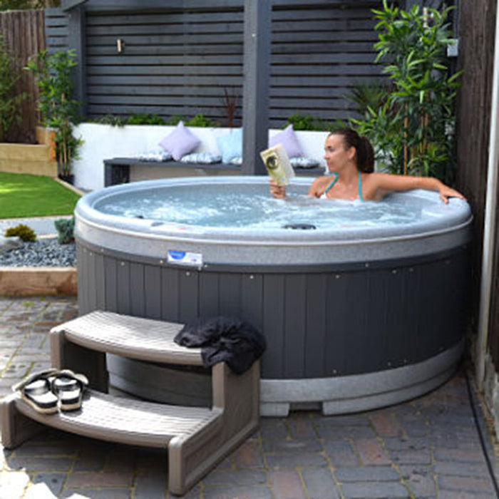 Woman relaxing in Rotospa Orbis Hot Tub reading a book in a stylish backyard garden setup.