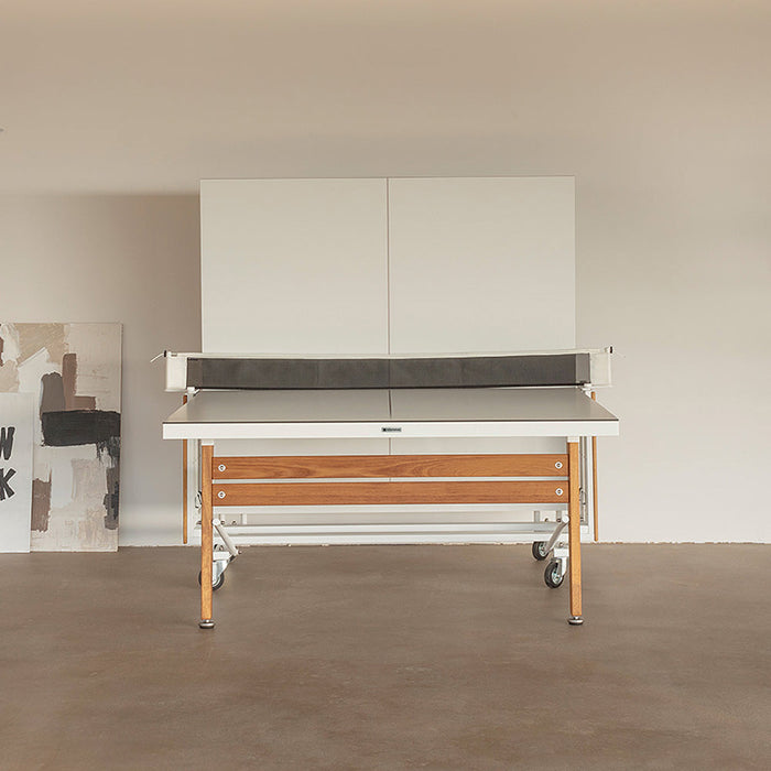Front view of RS Barcelona folding ping pong table with minimalist white top, wooden legs and central net system in a modern indoor space.