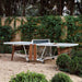 Outdoor folding ping pong table by RS Barcelona with sleek white tabletop, wooden legs and integrated net set in a garden setting for modern outdoor table tennis play.