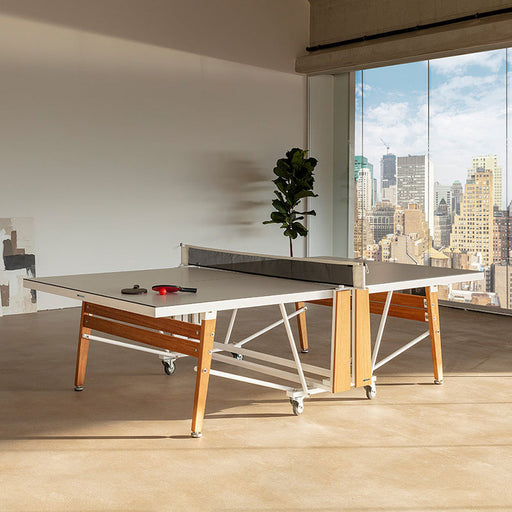 RS Barcelona folding table tennis table in stylish indoor space with city view, featuring white tabletop, wooden legs and modern net system.