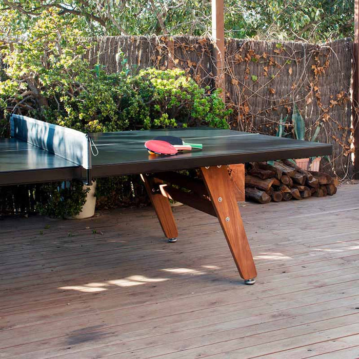 Outdoor RS Barcelona ping pong table with black surface and wooden legs on a garden deck surrounded by greenery and natural textures.