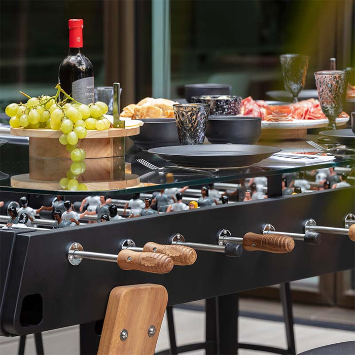 Close-up of RS Barcelona RS Max dining football table with glass top, wooden handles and players beneath, elegant food and wine presentation on multifunctional game dining table