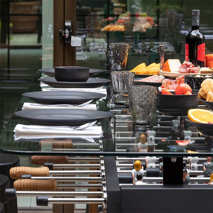 Side view of RS Barcelona RS Max dining football table with glass top, black plates and glassware, visible foosball rods and players beneath, modern indoor dining and gaming table