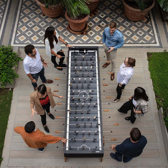 Overhead view of RS Barcelona RS Max football table with players gathered around in outdoor patio, modern foosball table with wooden handles and sleek black design for social gaming.