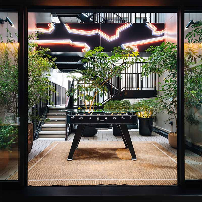 Black RS Barcelona RS2 dining football table in stylish indoor garden setting with glass top, blending foosball gameplay and contemporary dining furniture design.