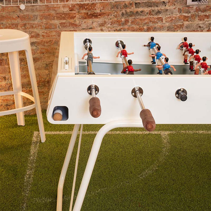 RS Barcelona RS3 football table in white finish with wooden handles, close-up of foosball players and rods on indoor turf-style flooring, modern compact game table design.