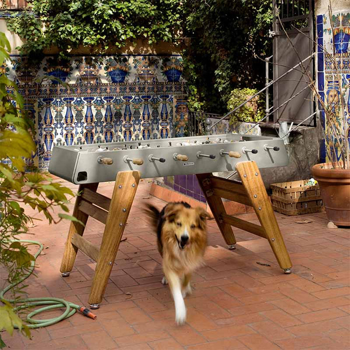 RS Barcelona RS3 Wood football table in outdoor courtyard with dog walking past, durable outdoor foosball table with wooden legs and modern neutral finish.
