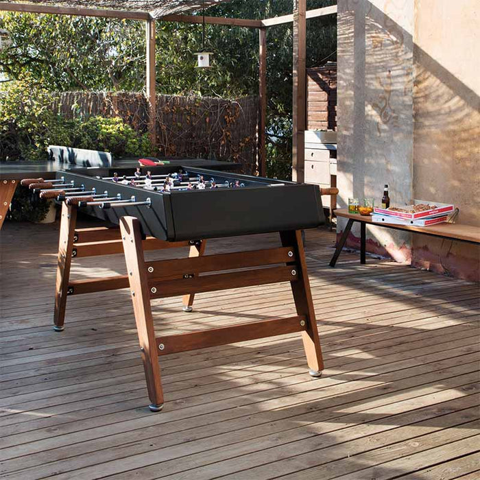 RS Barcelona RS3 Wood football table in sunlit outdoor terrace with wooden flooring and pergola, black foosball table with wooden legs in relaxed garden setting.