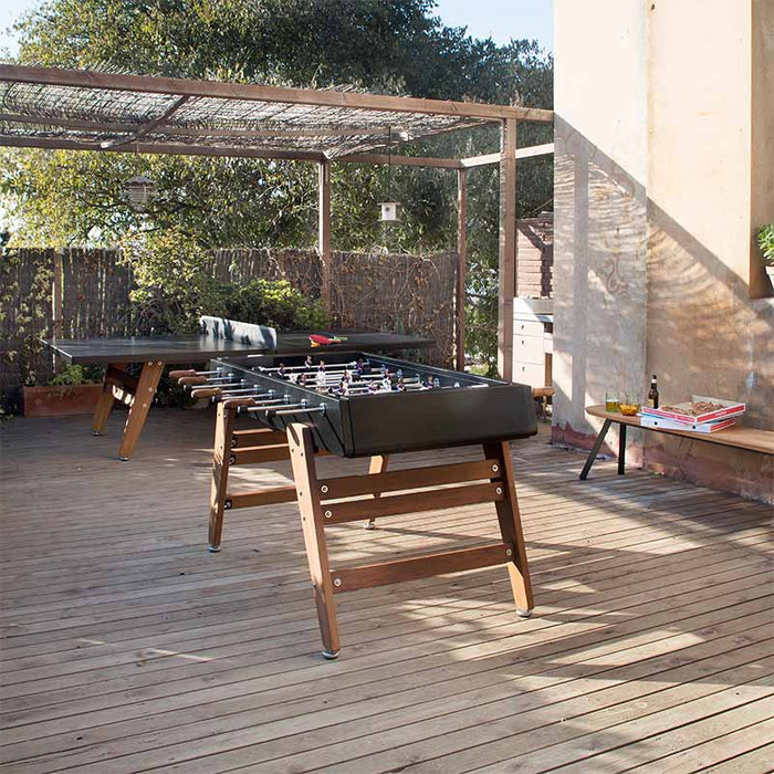 RS Barcelona RS3 Wood football table on outdoor wooden terrace with table tennis table and casual setup, modern foosball table with wooden legs in relaxed lifestyle setting.