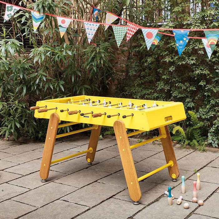 RS Barcelona RS4 Home football table in yellow with wooden legs placed outdoors in a garden setting, modern designer foosball table.