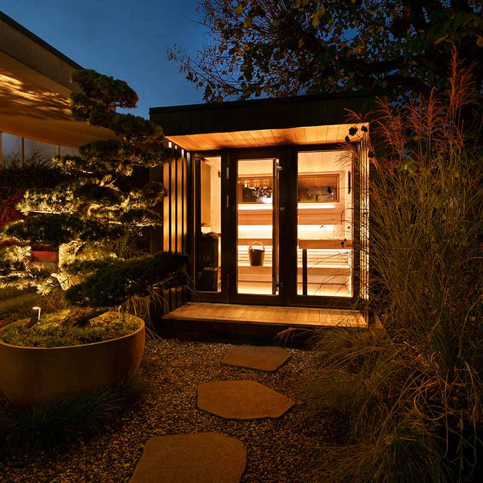 The Saunum AirCube Terrace Outdoor Sauna softly illuminated in a nighttime garden, blending into the tranquil outdoor setting with its warm interior lighting.