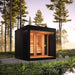 The Saunum AirCube Terrace Outdoor Sauna in a natural forest setting, featuring sleek black Thermo-Spruce cladding and large glass doors, blending into the surrounding greenery.