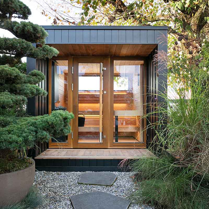 The Saunum AirCube Terrace Outdoor Sauna set on a patio, surrounded by outdoor greenery, with a stylish black exterior and wooden interior visible.