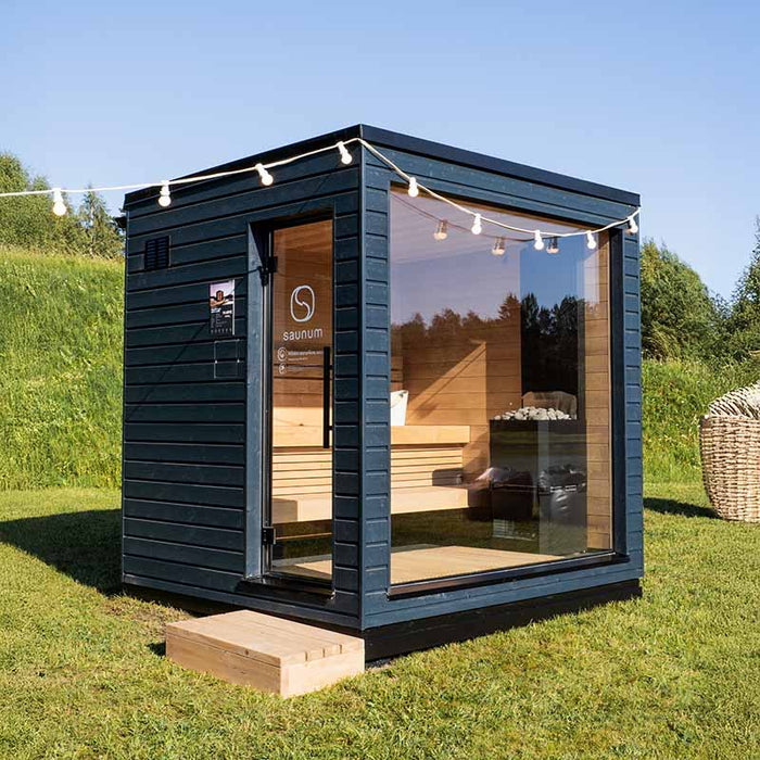 Saunum Classic Outdoor Sauna for 4 Persons, nestled in a natural setting, showcasing the Thermo-Spruce exterior and large glass panels.