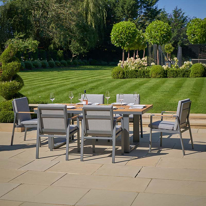 A view of the Stockholm 6 Seater Outdoor Dining Set arranged in a spacious garden, showcasing its modern design with grey cushions and teak wood accents.