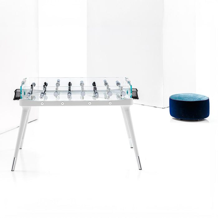 The elegant foosball table displayed in a minimalistic studio, emphasising its clear glass surface, white frame, and modern design. A blue pouffe is placed beside it.