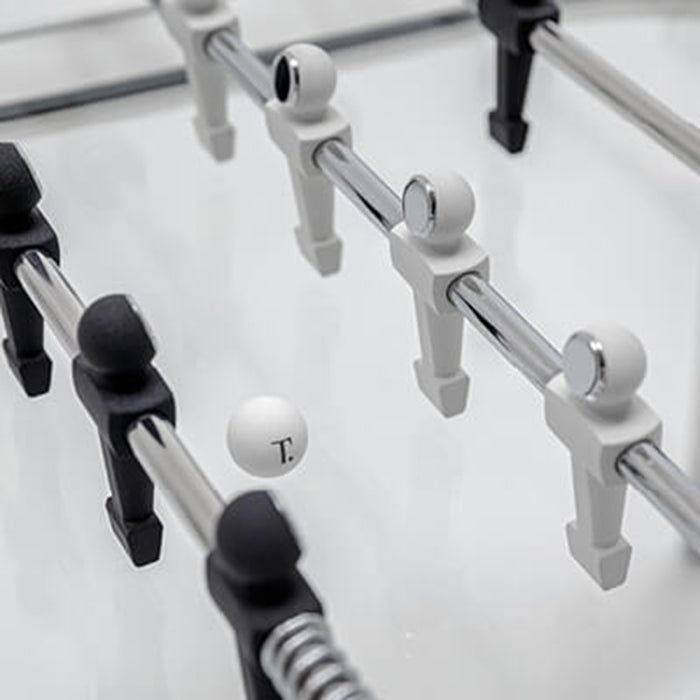 A detailed view of the white foosball used in the table, marked with a small 'T', lying on the clear glass playing surface.