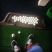 Top-down view of a golfer's shoes beside the True Strike golf mat with ball tray.