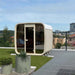 Side angle of the Ula Classic 6-person outdoor sauna in whitewashed finish on a rooftop garden, showcasing its modern design and serene placement.