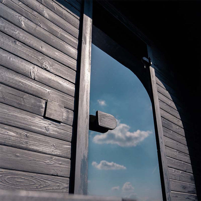 Close-up of the Ula Grand 6-Person Outdoor Sauna in Ebony finish with reflective glass door under a blue sky