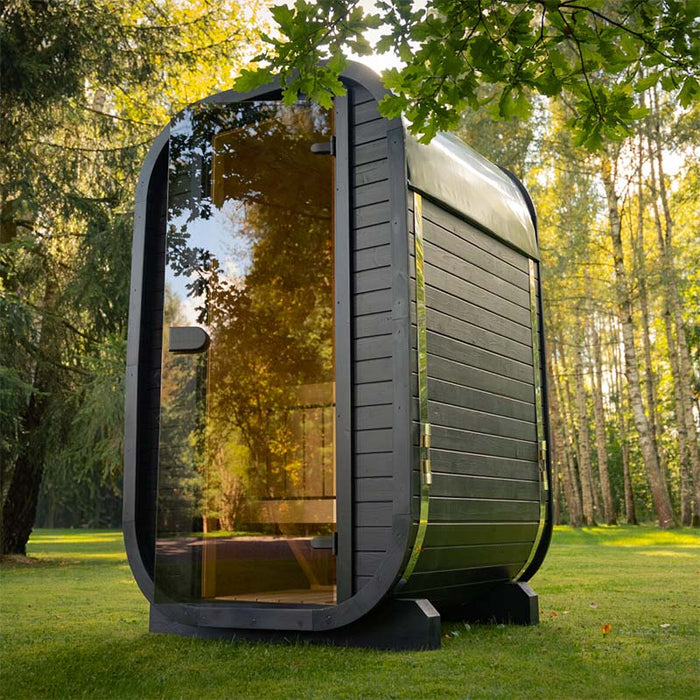 Angled view of the black Ula Micro 2-person outdoor sauna with its tinted glass door, blending into a natural woodland setting for outdoor relaxation.
