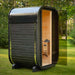 Black Ula Micro 2-person outdoor sauna with its glass door open, revealing a warm timber interior and Harvia heater, positioned in a peaceful garden setting.