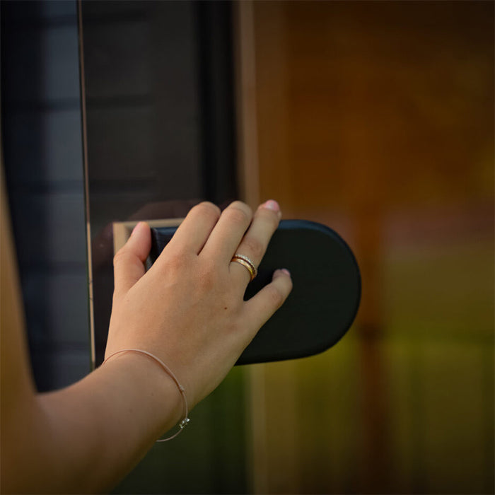 Close-up of a hand opening the tinted glass door of the Ula Micro 2-person outdoor sauna, showcasing the modern black handle and refined exterior finish.
