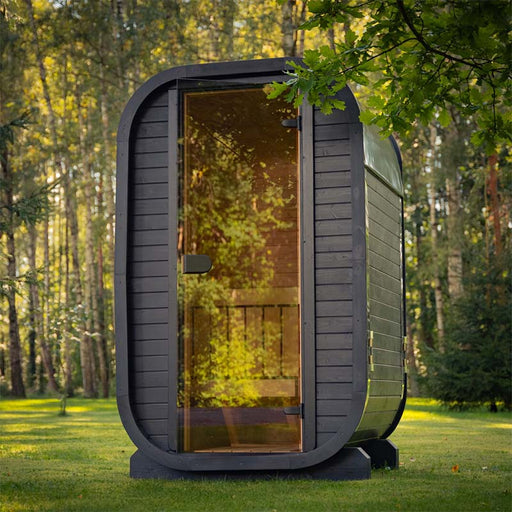 Front view of the Ula Micro 2-person outdoor sauna with tinted glass door and sleek black exterior, placed among tall trees for a serene outdoor wellness retreat.