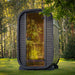 Front view of the Ula Micro 2-person outdoor sauna with tinted glass door and sleek black exterior, placed among tall trees for a serene outdoor wellness retreat.
