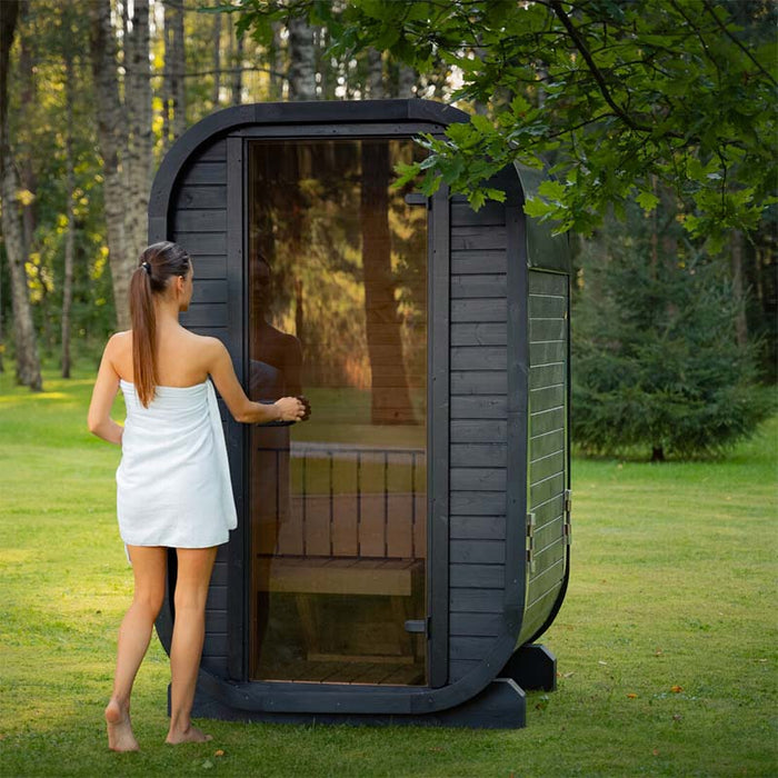 Person wrapped in a towel opening the tinted glass door of the Ula Micro 2-person outdoor sauna, set within a peaceful green garden environment.