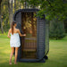 Person wrapped in a towel opening the tinted glass door of the Ula Micro 2-person outdoor sauna, set within a peaceful green garden environment.