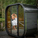Exterior front view of the black Ula Mini outdoor sauna with couple visible through the glass door.