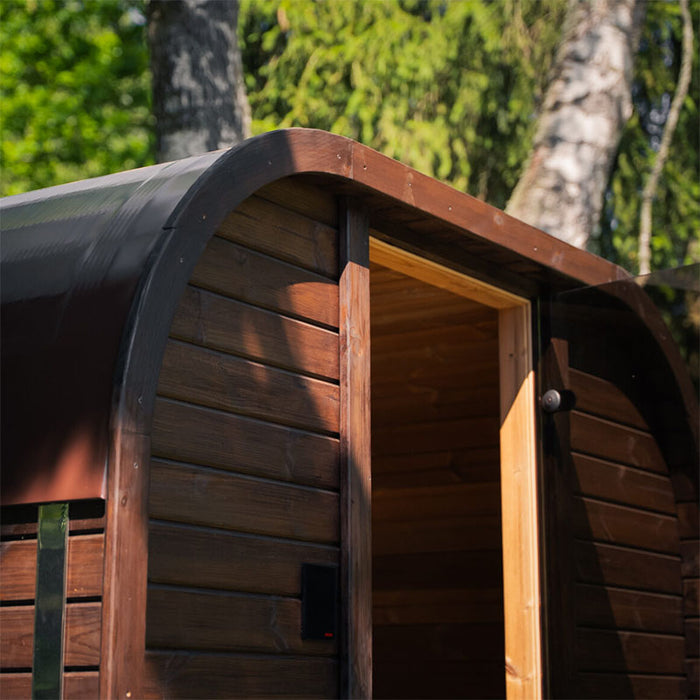 Angled side view of the Ula Mini sauna showing curved roof and smooth timber panelling against lush greenery.