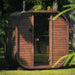 Ula Mini 2-person outdoor sauna with a rounded wooden exterior, positioned in a tranquil garden surrounded by trees.