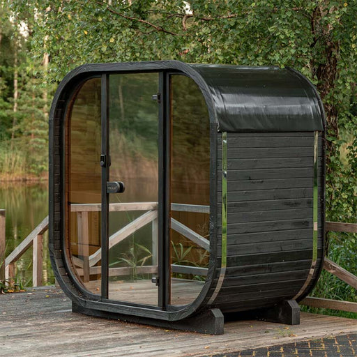 Couple walking towards the Ula Mini 2-person outdoor sauna positioned by a serene lakeside deck.