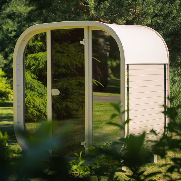 White Ula Mini outdoor sauna in a garden setting, surrounded by greenery with sleek glass door and curved design.