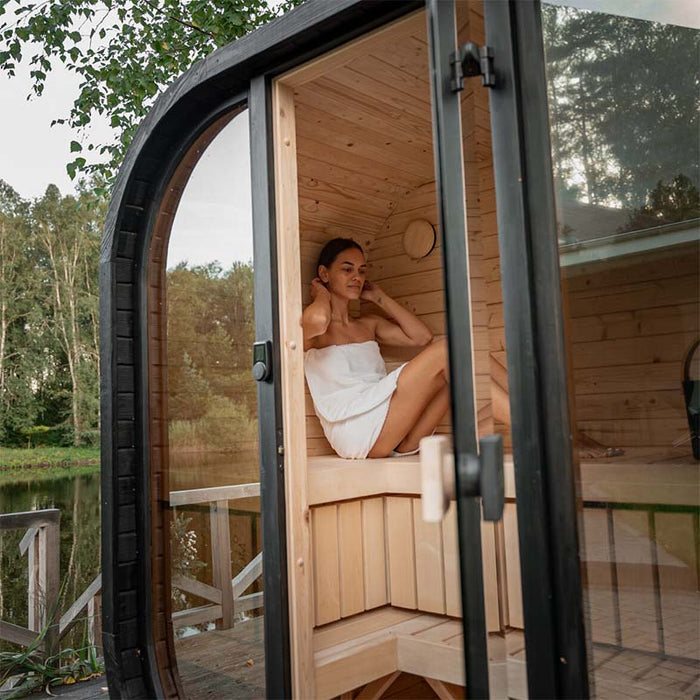 Woman relaxing inside the Ula Mini 2-person outdoor sauna, set beside a peaceful lakeside deck.