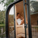 Woman relaxing inside the Ula Mini 2-person outdoor sauna, set beside a peaceful lakeside deck.