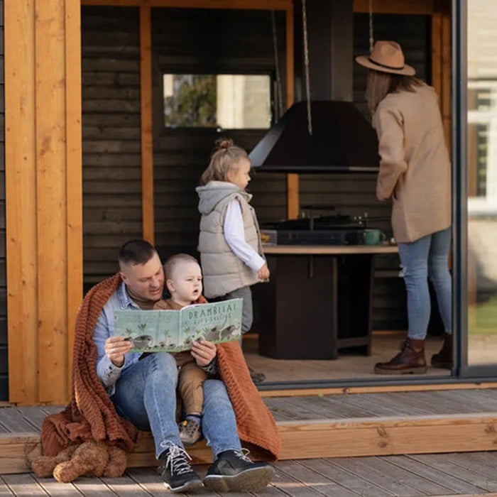 A father reading to his children outside the Garden Cube 3 x 8m, with a warm blanket and the barbecue area in the background, showcasing family-friendly design and cosy moments.