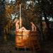  A couple relaxing in a 1.9m thermowood hot tub with a chimney, set in a peaceful woodland area.