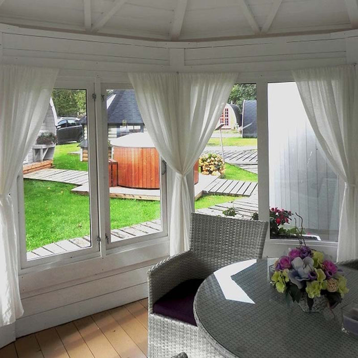 Scenic interior view of the Pavilion 10m² Sun showcasing white curtains, wicker furniture, and a beautiful garden setting visible through the glass walls.