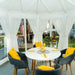 Bright interior of the Pavilion with Large Windows 10m², showcasing a stylish round dining table with yellow-accented chairs, surrounded by floor-to-ceiling glass windows and white curtains.