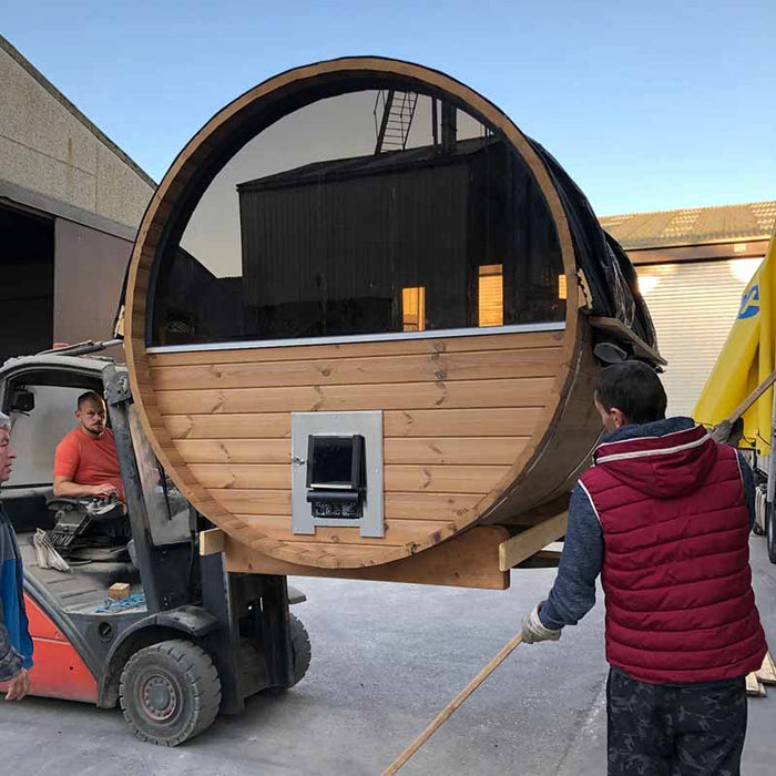 A forklift moving a traditional sauna barrel with workers guiding it.