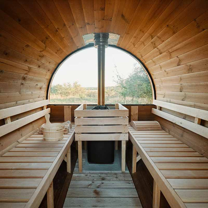 Interior of a Traditional Sauna Barrel 1.9 x 3m, showcasing wooden benches and a central sauna heater with a chimney, set against windows revealing an outdoor view.