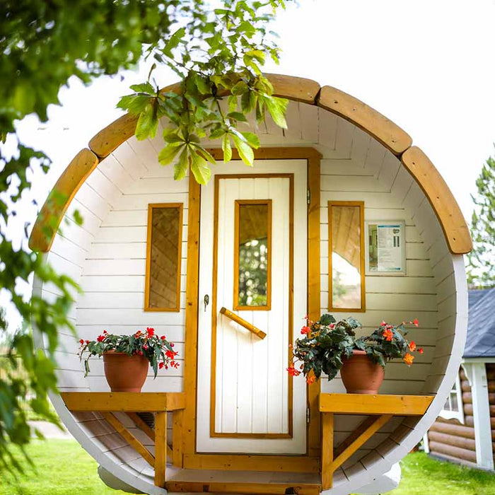 Traditional Sauna Barrel 1.9 x 3m, a round wooden sauna with a front door featuring glass panels, and flower pots on the porch, set in a lush garden