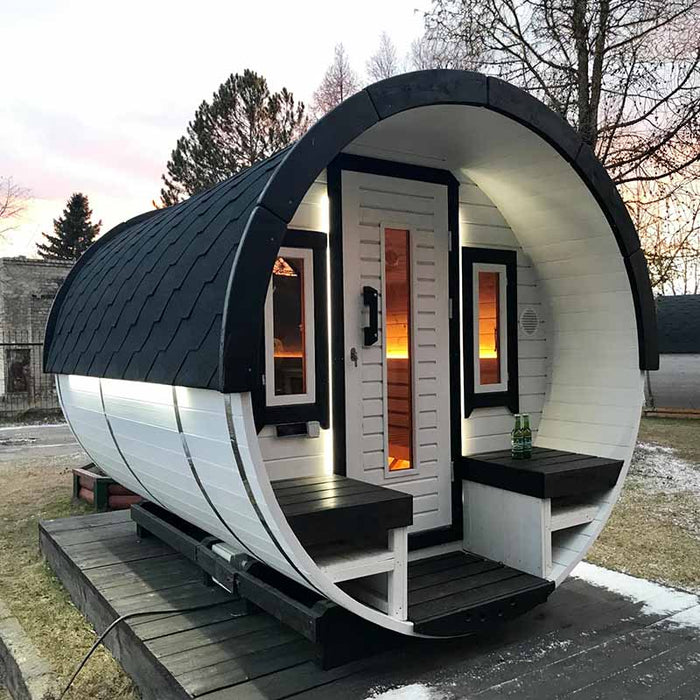 Traditional Sauna Barrel with open front door, interior benches, and warm lighting, set on a platform amidst grass and trees at dusk.